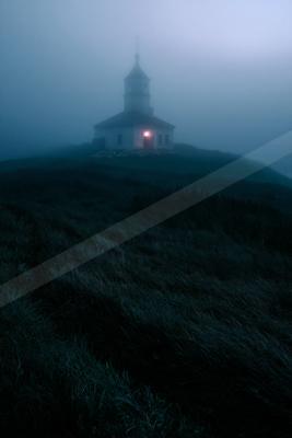 A small white church with a lit window and steeple stands on a foggy, grassy hill at night, surrounded by thick mist and dark vegetation.