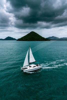 A white catamaran sailboat with full white sails glides across turquoise water, leaving a white wake, with a large forested island and dark storm clouds in the 
