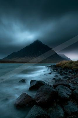 A dramatic mountain peak rises in the distance behind a misty lake with smooth water flowing around dark rocks in the foreground under heavy storm clouds.