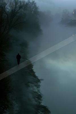 A lone silhouetted figure in a long coat walks along a snowy path beside a misty river, surrounded by bare trees and thick fog under dim blue light.