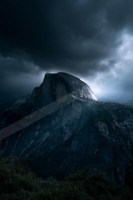 A massive granite mountain peak rises beneath heavy, dark storm clouds, with a beam of light breaking through on the right side, creating a moody and atmospheri