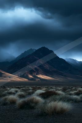 A rugged mountain peak rises against a sky filled with dark, heavy storm clouds, while dry desert shrubs cover the rocky foreground terrain.