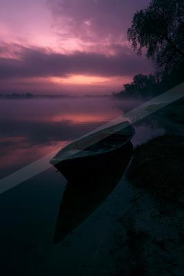 A wooden rowboat rests on the misty shore of a calm lake under a dramatic purple and pink sunset sky, with its reflection visible in the still water.