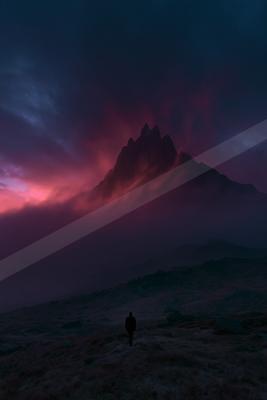 A lone person stands on a rocky moor looking toward a jagged mountain peak illuminated by intense red and purple clouds at dusk.