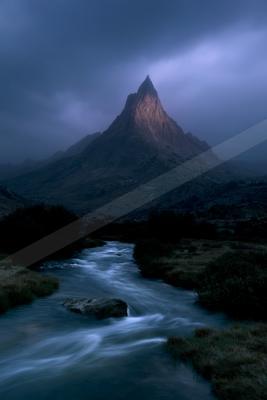 A sharp rocky mountain peak illuminated against dark stormy clouds, with a fast-flowing river in the foreground winding through a rugged valley with bushes and 