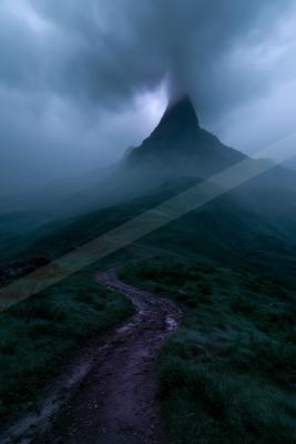 A winding dirt path leads through green hills toward a sharp, mist-covered mountain peak under a dark, cloudy sky.