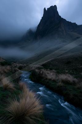 A fast-flowing stream winds through tufts of golden grass toward a towering, jagged mountain peak shrouded in thick fog and low clouds under a moody sky.