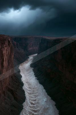 A powerful river flows through a deep, rugged canyon under dark, stormy clouds with dramatic light breaking through.
