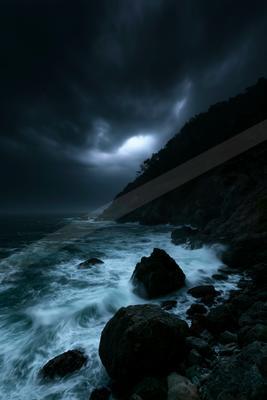Powerful ocean waves with white foam crash against dark jagged rocks on a rugged coastline beneath a moody, overcast stormy sky.