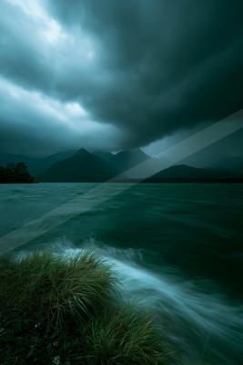 A dramatic stormy sky with dark clouds hangs over a mountain lake, where turbulent water meets a grassy rocky shore in the foreground.
