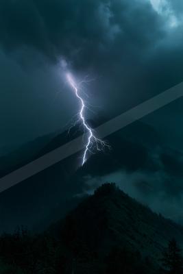 A bright white lightning bolt strikes a mountain peak during a dark, dramatic thunderstorm with heavy clouds and mist.