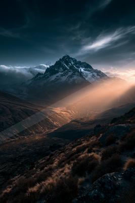 A majestic snow-capped mountain rises above a rugged valley under a moody sky filled with clouds, illuminated by the warm glow of sunset.