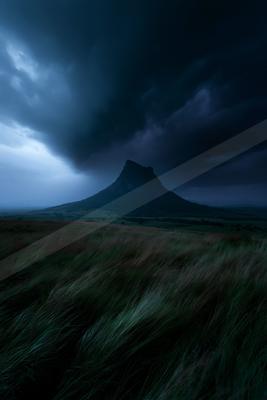 Dark storm clouds gather over a solitary mountain peak in a vast grassy plain, with wind-swept grass in the foreground.