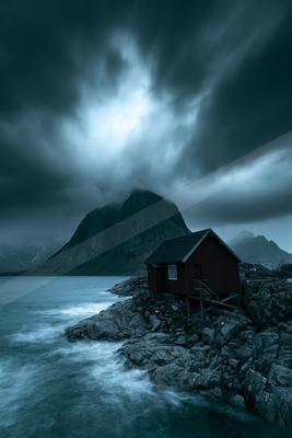 A red wooden cabin stands on a rocky shoreline next to a turbulent sea, with a large mountain partially obscured by dramatic, dark storm clouds in the backgroun