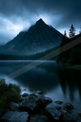A calm mountain lake reflects a large rocky peak partially shrouded in mist under a dark, cloudy sky, with foreground rocks and coniferous trees on the right.