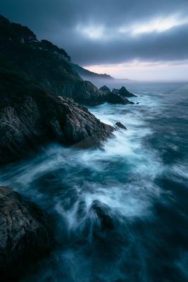Powerful ocean waves crash against rugged cliffs under a moody, overcast sky. The scene captures the raw energy of nature as mist rolls in from the horizon.