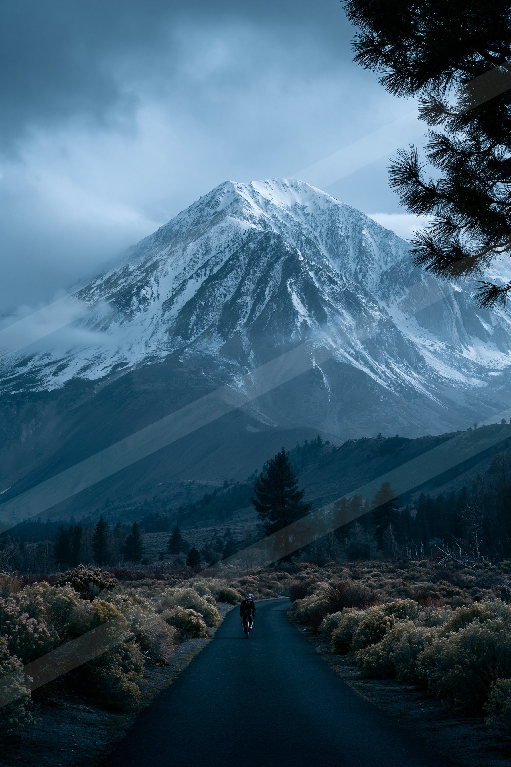 A lone cyclist rides along a dark road leading towards a towering snow-covered mountain under an overcast sky.