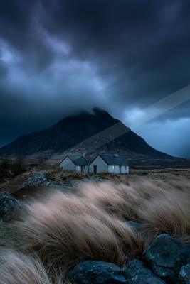 Two traditional stone cottages stand in a windswept moorland foreground with tall grass blowing in the wind, set against a large dark mountain partially shroude