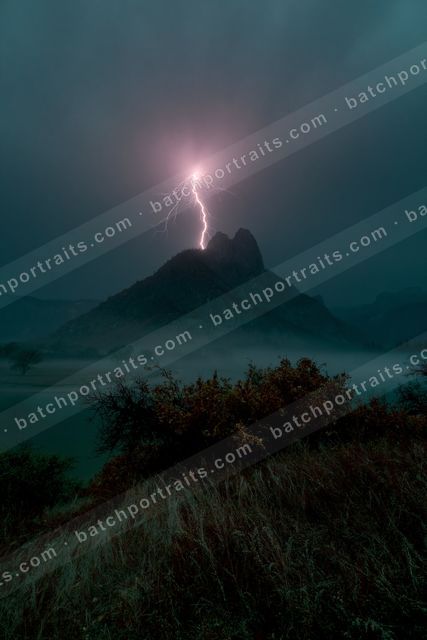 A powerful lightning bolt strikes the peak of a mountain shrouded in mist, with silhouetted trees and grasses in the foreground.