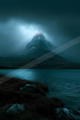 A sharp, snow-capped mountain peak rises through thick fog and low clouds above a calm alpine lake, with rocky grassy shore in the foreground under a dark moody