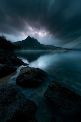 Dark rocky shoreline with large boulders in a calm mountain lake under a dramatic stormy sky with breaking light over a distant peak.