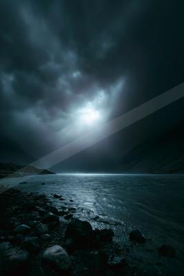 Moody atmospheric scene of a dark lake under heavy storm clouds with a bright break in the center, rocky shoreline in foreground