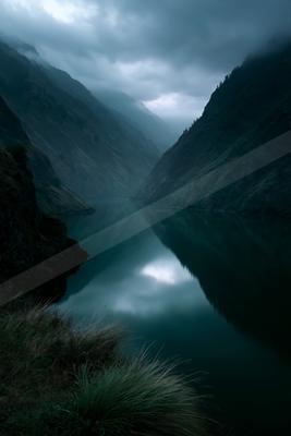 A calm mountain lake reflects the steep, forested slopes and overcast sky within a misty canyon, with tall grass in the foreground.