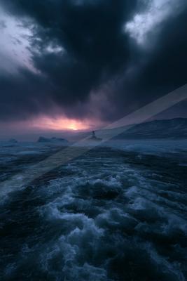A naval warship sails through choppy dark waters filled with ice floes under a dramatic stormy sky with a glowing sunset on the horizon.