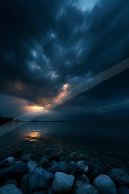 Moody dark storm clouds illuminated by a break of golden light at the horizon, reflecting on a calm lake with a foreground of rounded stones.