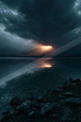 Moody landscape of a calm mountain lake reflecting a dramatic sunset breaking through dark storm clouds, with rocky shoreline in the foreground.