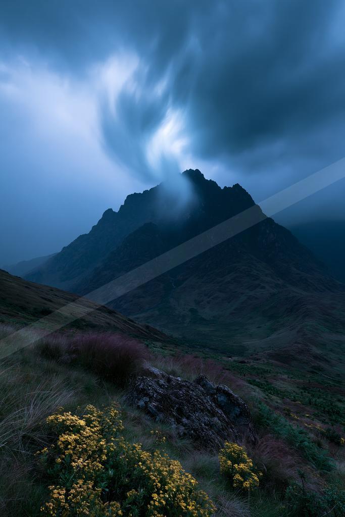 A rugged mountain peak rises into swirling blue and white clouds in a dramatic stormy sky, with yellow wildflowers and rocky terrain in the foreground.