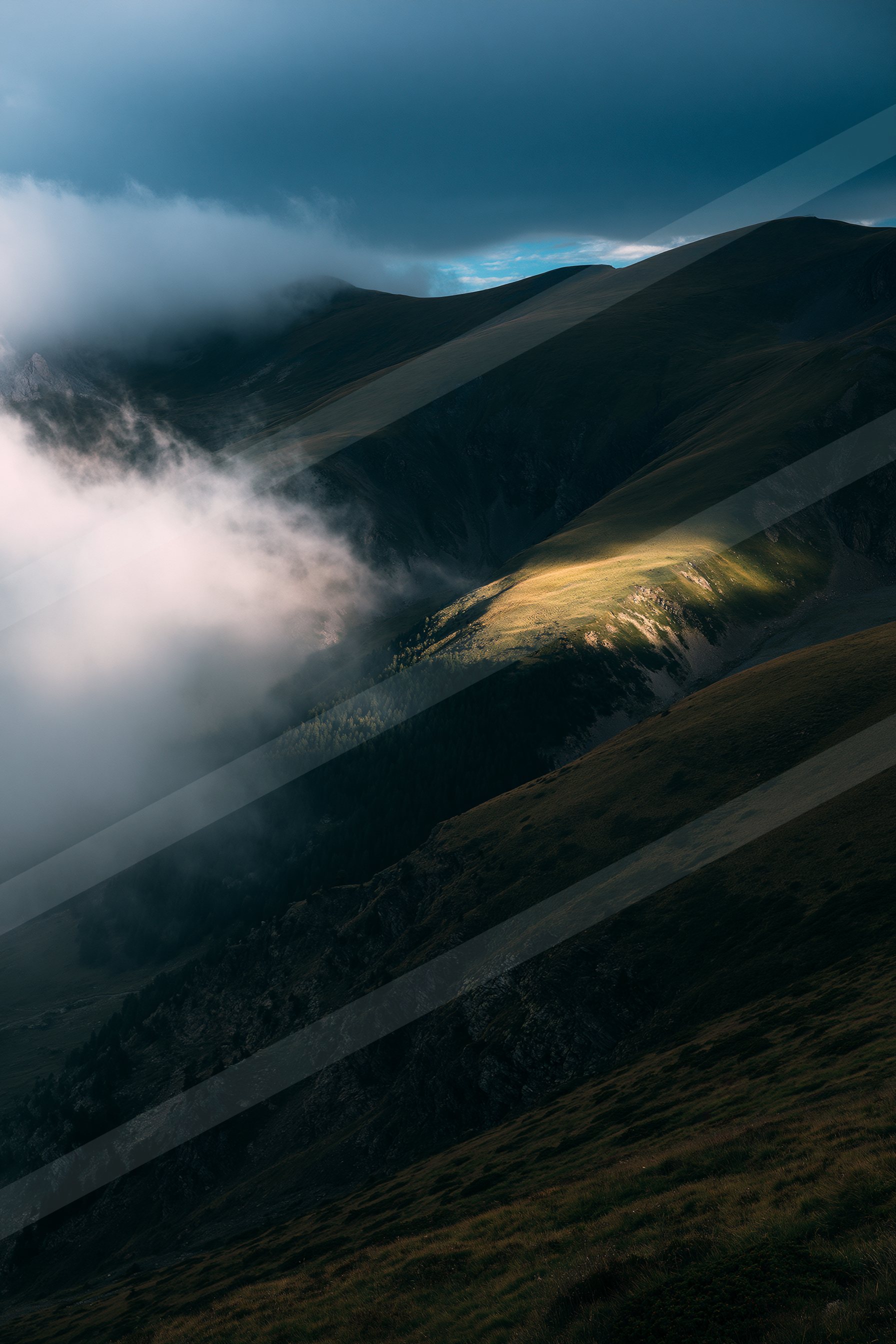 A mountainous landscape featuring a sunlit peak surrounded by dark, forested slopes and dramatic clouds in the sky.