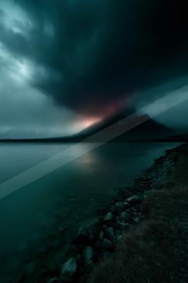 Moody landscape with dark storm clouds hovering over a mountain range, reflecting in a calm lake with a rocky shoreline in the foreground.