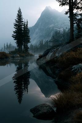 A calm mountain lake perfectly reflects tall pine trees and a rocky peak shrouded in mist, with boulders and shrubs in the foreground.