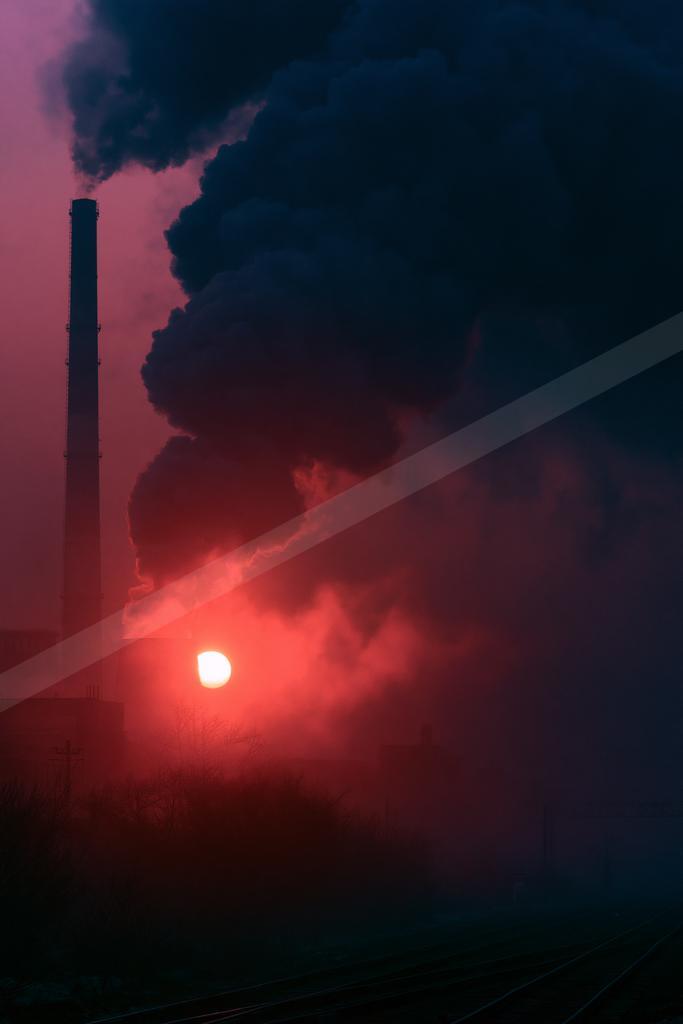 A tall industrial smokestack emits thick dark smoke against a vivid red and purple sunset sky, with the sun glowing brightly near the horizon and railway tracks