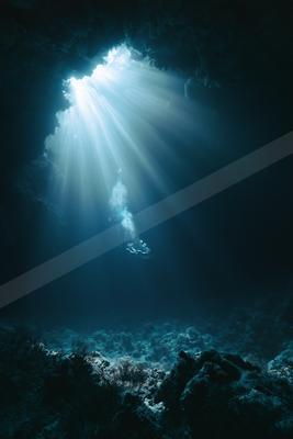 A scuba diver swims through dramatic crepuscular rays of sunlight penetrating a dark underwater cave, illuminating bubbles and rocky formations.