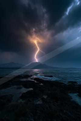 A powerful lightning bolt strikes a distant mountain peak during a stormy night, illuminating dark clouds above a rocky shoreline and choppy sea.