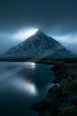 A majestic snow-covered mountain peak rises under dramatic dark clouds, with bright light breaking through at the summit, its reflection visible on the smooth s