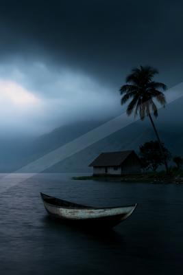 A weathered wooden boat floats on a calm lake in the foreground, with a small house and tall palm tree on a distant shore under a dark, cloudy sky with mountain