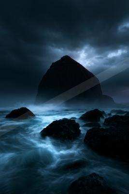 A large dark sea stack rises from turbulent blue water under a brooding stormy sky, with waves crashing against foreground rocks in a moody coastal scene.