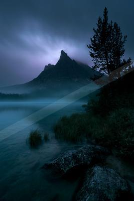 A serene mountain lake shrouded in mist under a dramatic twilight sky, with a tall pine tree on the right shore and large rocks in the foreground.