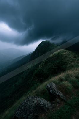 Dark storm clouds loom over a lush green mountain ridge with rocky foreground and mist-filled valleys.