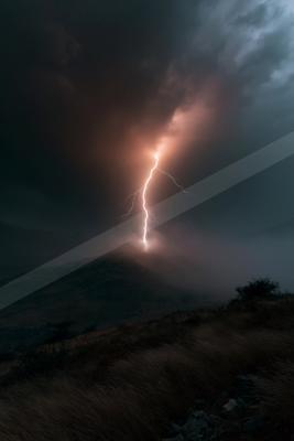 A bright white lightning bolt strikes the peak of a dark hill under a turbulent, cloudy sky, illuminating the surrounding mist and landscape.