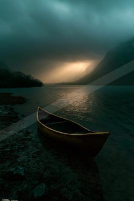 A yellow canoe rests on the rocky shore of a calm mountain lake under a dramatic, misty sky with light breaking through the clouds.
