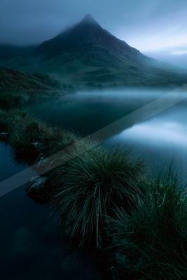A serene mountain lake surrounded by grassy banks and rocks, with thick fog hovering over the water and a large peak rising into the cloudy sky.