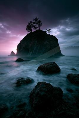 A large rocky sea stack topped with silhouetted trees rises from a misty ocean under a dramatic purple and pink twilight sky, with smooth water flowing around d
