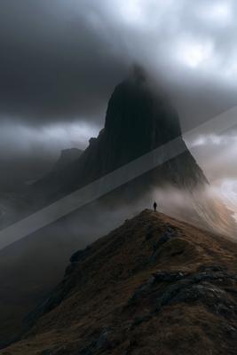 A solitary person stands on a rocky mountain ridge overlooking a massive, fog-shrouded peak under a brooding, cloudy sky.