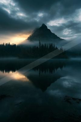 A dark mountain peak rises above a tranquil lake, shrouded in mist and reflected perfectly on the still water's surface. The sky is filled with moody clouds ill