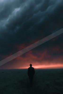 A solitary man in a dark coat walks away from the viewer across a grassy field toward a dramatic sky filled with dark storm clouds and a glowing red-orange hori
