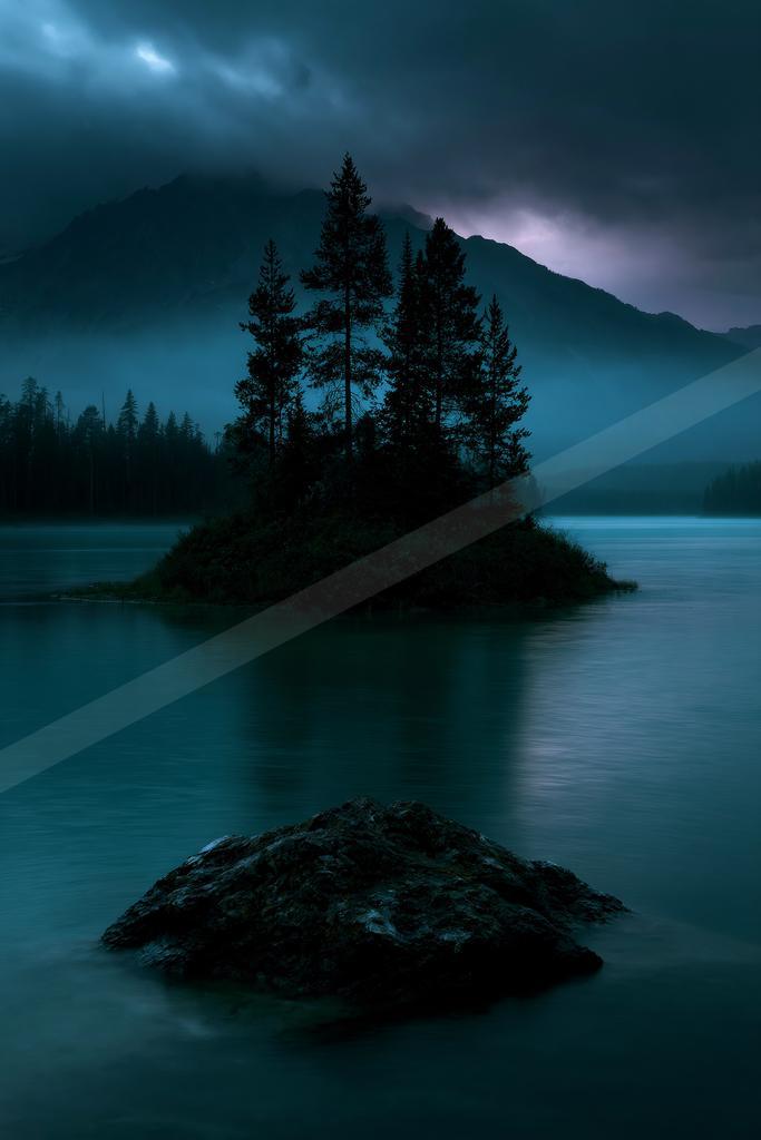 A small rocky island covered in tall evergreen trees sits in the middle of a calm mountain lake, with a large dark rock in the foreground and misty mountains un